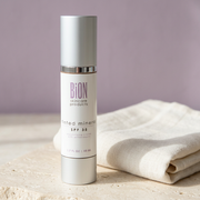 BiON Tinted Mineral SPF 35 silver bottle on pale stone beside ivory linen and lilac backdrop